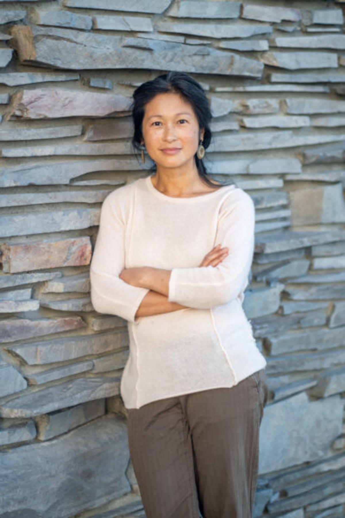 Linda Thai color portrait. Linda is a Vietnamese woman who is standing and leaning against a stone wall. Her arms are crossed in front of her and is smiling.